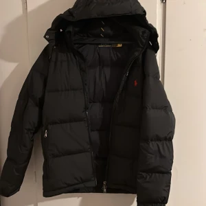 Svart dunjacka från Polo Ralph Lauren - Snygg svart dunjacka från Polo Ralph Lauren med röd broderad logga på bröstet. Jackan har en praktisk dragkedja och en justerbar huva. Perfekt för kyliga dagar.