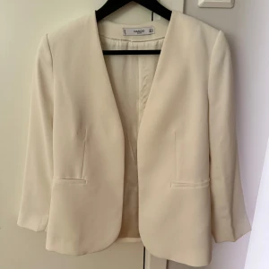 Beige kavaj från Mango - Säljer en stilren äggskalsfärg kavaj från Mango i storlek M. Kavajen har en elegant och enkel design utan knappar, vilket ger den en modern look. Perfekt för både vardag och festliga tillfällen. Den är i fint skick och redo för en ny ägare!