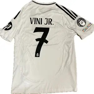 Säljer en fotbollströja från Real Madrid med Vini Jr. #7 på ryggen. Tröjan är en reprint. Storlek: S. Ansvarar ej för postens slarv. Endast handtvätt gäller, gnugga ej på blöta prints.