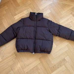 Brun pufferjacka från Weekday - Säljer en snygg brun pufferjacka från Weekday i storlek M. Jackan har en hög krage och stängs med dragkedja. Perfekt för kyliga dagar och ger en cool look med sin puffiga stil. Den är i bra skick och redo för en ny ägare! Saknar tillhörande luva.