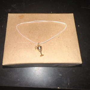 Armband med guldfärgad berlock - Säljer ett stilrent armband med en tunn kedja och en charmig guldfärgad berlock i form av ett vinglas. Perfekt för festliga tillfällen eller som en rolig detalj till vardagsoutfiten.