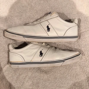 Vita sneakers från Polo Ralph Lauren - Säljer ett par vita sneakers från Polo Ralph Lauren. Skorna har en klassisk design med snörning och den ikoniska Polo-loggan på sidan. De är tillverkade i skinn med en gummisula, vilket gör dem både stiliga och bekväma. Perfekta för vardagsbruk!