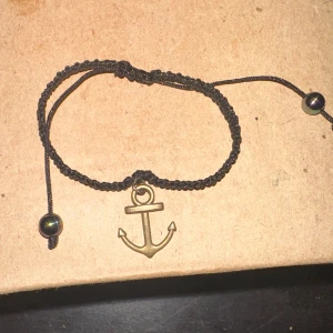 Svart armband med ankare - Säljer ett snyggt svart armband med ett guldigt ankare som hänge. Armbandet är justerbart med små pärlor i ändarna. Perfekt för en avslappnad stil eller som en present till någon speciell!