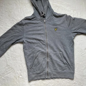 Grå hoodie från Lyle & Scott - Säljer en snygg grå hoodie från Lyle & Scott. Den har en dragkedja framtill och en broderad gul logga på bröstet. Perfekt för en avslappnad stil och passar till alla tillfällen. Den är i okej skick och superbekväm! Är 180 och väger 75kg