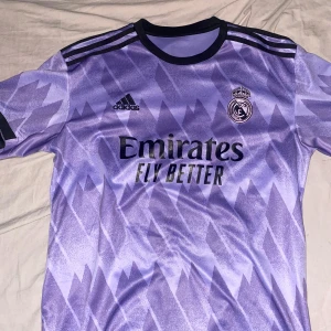 Lila fotbollströja från Adidas - Snygg lila fotbollströja från Adidas med Real Madrid-logga och 'Emirates Fly Better' tryck. Tröjan har ett unikt mönster och korta ärmar, perfekt för fotbollsfans.