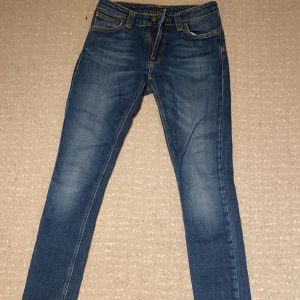 Blåa nudie jeans - Snygga blå jeans från Nudie Jeans med klassisk femficksdesign och orange sömmar. De har en slim passform och är perfekta för en stilren look. Märkeslogga på bakfickan och läderpatch i midjan. Skick: 10/10