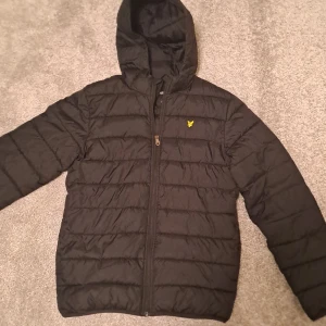 Svart dunjacka från Lyle & Scott Junior - Snygg svart dunjacka från Lyle & Scott Junior med huva och dragkedja. Jackan har ett quiltat mönster och en liten gul logotyp på bröstet. Perfekt för kyligare dagar.Den är köpt från kids brandstore för 1100 och är i nyskick 