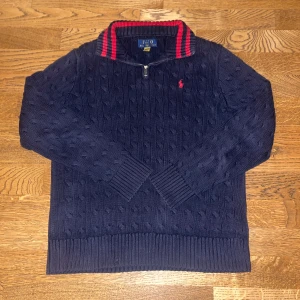 Mörkblå kabelstickad tröja från Ralph Lauren - Säljer en snygg mörkblå kabelstickad tröja från Ralph Lauren med dragkedja och röd broderad logga på bröstet. Tröjan har en ribbad krage med röda ränder och långa ärmar. Perfekt för kyliga dagar! Tröjan har även ett ytterst litet hål längst ner som är igensytt. Men när man har på sig tröjan så syns det inte om man inte kollar på väldigt nära håll. Pris kan diskuteras
