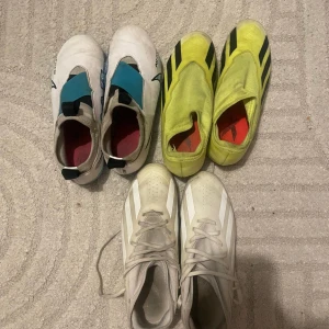 Tre par fotbollsskor - Säljer tre par fotbollsskor i olika färger och stilar. Ett par är vita med blå detaljer, ett par är gula med svarta ränder, och det tredje paret är vitt med snörning och diskreta ränder.