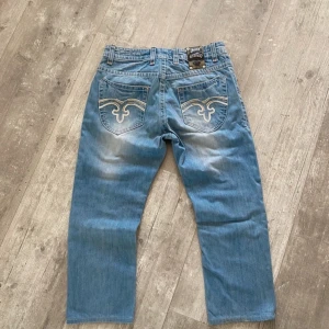 Rock Revival Jeans  - Snygga blå jeans från Rock Revival med unika broderade detaljer på bakfickorna. Jeansen har en klassisk femficksdesign och en knappgylf. Pris går att diskutera vid snabbköp! 