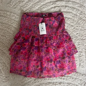 Blommig rosa kjol från H&M - Säljer en söt blommig rosa kjol från H&M med volanger. Kjolen har en elastisk midja och är perfekt för sommarens varma dagar. Den är kort och har ett färgglatt mönster i olika nyanser av rosa och lila.