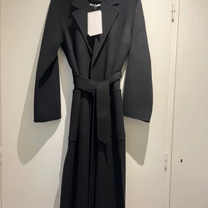 Svart kappa från Ivy Oak - Elegant svart kappa från Ivy Oak i 100% ull. Den har en klassisk design med knytskärp i midjan och långa ärmar. Perfekt för kyligare dagar.