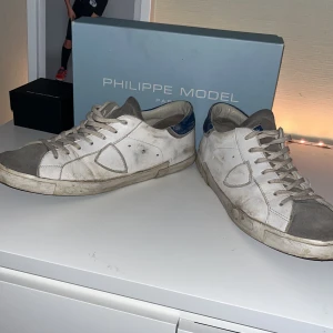 Vita sneakers från Philippe Model - Snygga vita sneakers från Philippe Model med grå detaljer och blå häl. Skorna har snörning och en klassisk design som passar perfekt till vardags. De är tillverkade i skinn och har en bekväm passform. Kasta in dem hos en sko makare för reparation så kan dem nog bli riktigt fräscha igen. Box ingår lågt pris pga skick