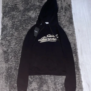 Svart hoodie från Saint Laurent - Snygg svart hoodie från Saint Laurent med logga i vitt på bröstet. Tröjan har en klassisk design med luva och en ficka, Väldigt bra kvalite och passar perfekt för allt.