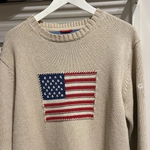 Beige stickad tröja från GANT - Säljer en snygg beige stickad tröja från GANT med ett amerikanskt flaggmotiv på framsidan. Tröjan har en rund halsringning och är tillverkad i 100% bomull. Perfekt för en avslappnad stil.
