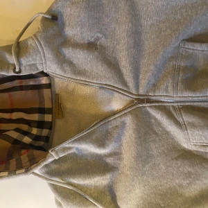 Grå hoodie med dragkedja - Snygg grå hoodie från Burberry med dragkedja och klassiskt rutigt mönster i huvan. Den har en broderad logga på bröstet och är perfekt för en avslappnad stil. Nypris 7700, mitt pris 599 