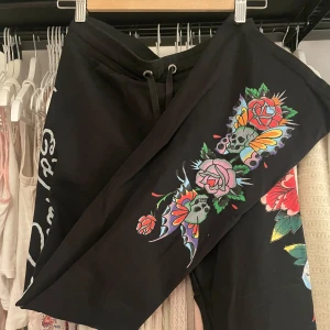 Svarta mjukisbyxor från Ed Hardy - Snygga svarta mjukisbyxor från Ed Hardy med färgglada blommiga och dödskallemotiv. Byxorna har snörning i midjan och tryck längs benen. Utsvändga och midrise. Använda några få gånger. 💓🍓