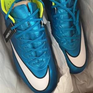 Blå Nike Mercurial fotbollsskor - Snygga blå Nike Mercurial vapor X fotbollsskor med vit swoosh och neongula detaljer. Skorna har snörning och en slitstark sula med dobbar för optimalt grepp på planen. Perfekta för fotbollsspelare som vill ha lätta skor tillsammans med stil och funktion.