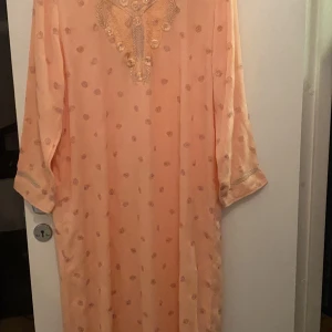 Persikofärgad klänning med broderi - Elegant persikofärgad klänning med broderade detaljer och diskret prickigt mönster. Klänningen har trekvartsärmar och en lös passform, perfekt för en avslappnad men stilfull look. Köpte den i Dubai för 999kr helt oanvänt 