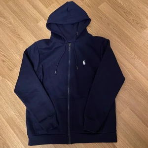 Mörkblå hoodie från Ralph Lauren - Säljer en stilren mörkblå hoodie från Ralph Lauren med dragkedja och det klassiska logotypbroderiet på bröstet. Tröjan har en justerbar huva och ribbade muddar vid ärmslut och nederkant. Perfekt för en avslappnad stil. Helt Oanvänd (F@jk)
