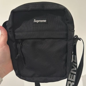 Svart axelväska från Supreme - Snygg svart axelväska från Supreme med justerbar axelrem och dragkedja. Perfekt för att bära med sig det viktigaste. Väskan har en stilren design med Supreme-loggan på framsidan.