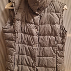 Grå  grön dunväst från Escada Sport - Snygg och stilren grå dunväst från Escada Sport. Västen har en quiltad design med knappar framtill och en hög krage för extra värme. Perfekt för kyligare dagar.