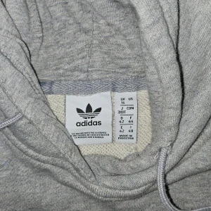 Grå hoodie från Adidas - Säljer en grå hoodie från Adidas med det klassiska logotyptrycket i vitt på framsidan. Tröjan har en stor ficka framtill och justerbar huva med dragsko. Perfekt för en avslappnad stil.