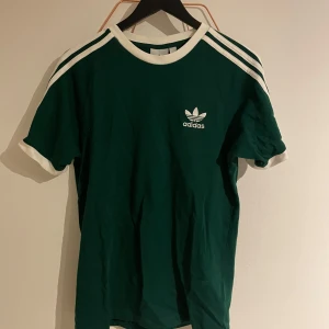 Grön t-shirt från Adidas - Säljer en grön t-shirt från Adidas med vita detaljer och klassiska ränder på axlarna. T-shirten har korta ärmar och en rund halsringning. Perfekt för en sportig look!