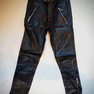 Svarta byxor av skinnimitation med dragkedjor - Snygga svarta byxor av skinnimitation med flera dragkedjor för en edgy look. Byxorna har en tight passform och är perfekta för att ge din outfit en cool touch. De är designade med detaljer som dragkedjor vid fickorna och bensluten.