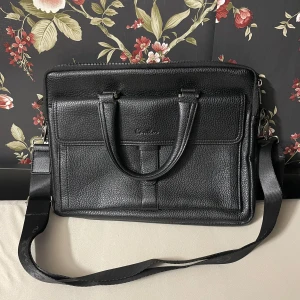 Svart datorväska från Cantlor - Snygg och praktisk svart datorväska. Väskan har både handtag och en justerbar axelrem för bekväm användning. Den har flera fack med dragkedja för enkel organisering av dina saker. Perfekt för både arbete och studier.