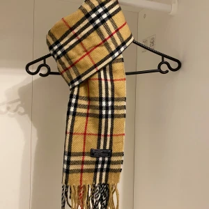 Burberry - Säljer nu denna Snygga halsduk från Burberry i beige. Tillverkad i 100% lammull material med fransar i ändarna. Perfekt för att hålla dig varm och stilren under kyliga dagar.