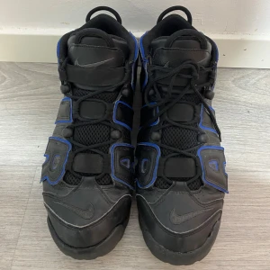 Nike Air More Uptempo i svart och blå - Säljer ett par Nike Air More Uptempo sneakers i svart med blå detaljer. Skorna har en iögonfallande design med stora bokstäver på sidan och snörning framtill. Perfekta för dig som vill ha en unik stil.