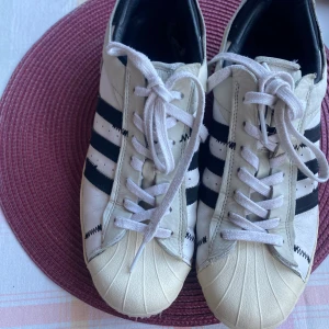Adidas Superstar sneakers - Klassiska Adidas Superstar sneakers i vitt med svarta ränder. Skorna har snörning och den ikoniska tåhättan i gummi. Perfekta för en stilren och sportig look. Säljes då dem är för stora för mig!