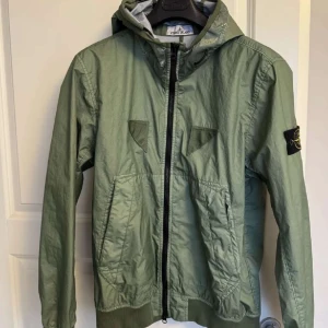 Grön vindjacka från Stone Island - Snygg grön vindjacka från Stone Island med huva och dragkedja. Jackan har en ikonisk patch på ärmen och praktiska fickor framtill. Perfekt för vår och höst. Skicket är som ny och självklart är den äkta. Originella pris 8000 ny pris 4000. Hjälper en vän sälja. Storleken är M men passar S.