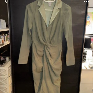 Olivgrön omlottklänning - Elegant olivgrön omlottklänning med långa ärmar och knytning i midjan. Klänningen har en stilren krage och är perfekt för en chic look.