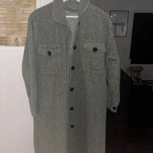 Grön lång kappa med knappar - Säljer en stilren grå kappa med långa ärmar och knappar framtill. Den har två stora fickor med knappar på bröstet. Tillverkad i Frankrike av 95% polyester och 5% elastan.