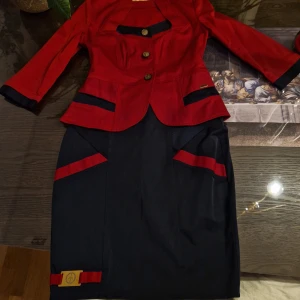 Röd och marinblå dräkt - Elegant dräkt i rött och marinblått med trekvartsärmad kavaj och knälång kjol. Kavajen har dekorativa knappar och kontrasterande detaljer i marinblått. Kjolen har matchande röda detaljer och en diskret logotyp vid fållen.