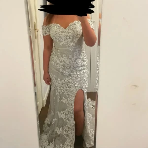 Vit spetsklänning med slits - Elegant vit långklänning med vacker spets och offshoulder-design. Klänningen har en djärv slits framtill och en figurnära passform som framhäver silhuetten.