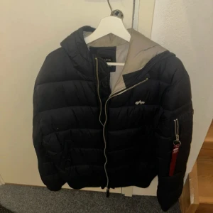 Svart pufferjacka från Alpha Industries - Säljer en svart pufferjacka från Alpha Industries med dragkedja och huva. Jackan har en cool detalj med en röd 'Remove Before Flight'-tag på ärmen. Perfekt för kyliga dagar. Använd och lite smutsig runt kragen och ett litet hål som ni kan se på bilden 