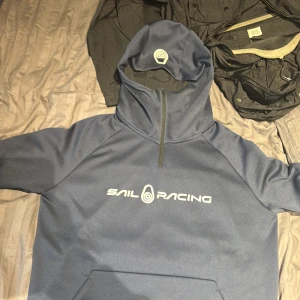 Blå hoodie från Sail Racing - Säljer en snygg blå hoodie från Sail Racing med dragkedja vid halsen och logotyp på bröstet. Perfekt för en sportig look eller en avslappnad dag. Hoodien har en praktisk ficka framtill.
