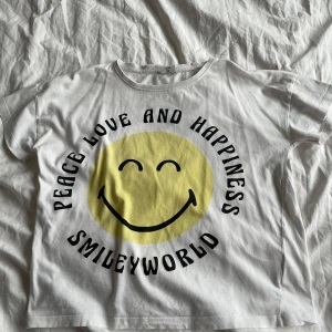 Vit t-shirt med smiley från SmileyWorld😀 - Vit t-shirt från SmileyWorld med ett stort gult smileytryck och texten 'Peace Love and Happiness'. Perfekt för en avslappnad stil❤️