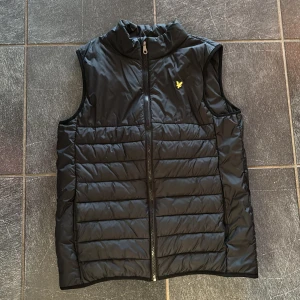 Svart  lyle scott dunväst med dragkedja - Snygg svart lyle Scott dunväst med dragkedja och broderad logga på bröstet. Perfekt för lager-på-lager under kyliga dagar. Västen har en stilren design och är lätt att matcha med olika outfits. Den är 14/15 år men passar bra mig då jag säljer för den blivit för kort, passar nog perfekt om du är c 170 