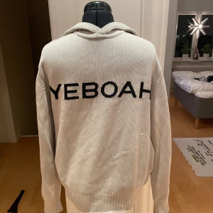 Cos X Yeboah hoodie - Snygg beige stickad hoodie med texten 'YEBOAH' i svart på framsidan och en svart stjärna på ryggen. Perfekt för en avslappnad stil med en unik touch.