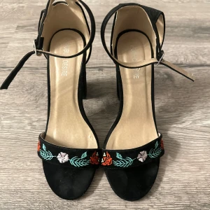 Svarta klackskor med broderade blommor - Eleganta svarta klackskor från Public Desire med vackra broderade blommor i rött, vitt och grönt. Skorna har en öppen tå och justerbar ankelrem för perfekt passform. Perfekta för att ge en färgglad touch till din outfit.