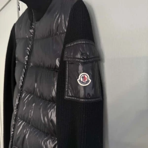 Svart dunjacka från Moncler - Snygg svart dunjacka från Moncler med stickade ärmar och dragkedja framtill. Jackan har en quiltad design och Moncler-logga på ärmen. Köpt i Paris, lite stor i storleken. Köpt för 790 euro, mycket bra skick. Kvittot finns digitalt. 