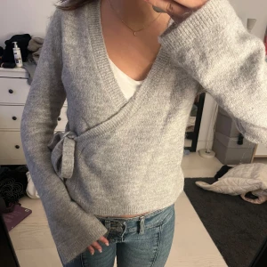 Grå omlottkofta - Mysig grå omlottkofta med knytband i midjan. Tröjan har långa ärmar och är perfekt för kyligare dagar. Den är tillverkad i ett mjukt material som ger en bekväm passform.