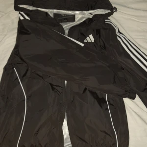 Svart vindjacka från Adidas - Snygg svart vindjacka från Adidas med vita ränder på ärmarna och dragkedja framtill. Jackan har en huva och är perfekt för blåsiga dagar. Den har en sportig stil med klassiska Adidas-detaljer fungerar perfekt till träningar utomhus när det är kallt. Helt ny passar dig mellan 155-165cm