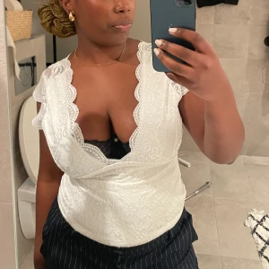 Vit spetsblus - Säljer en vacker vit spetsblus med djup v-ringning och fina spetsdetaljer. Blusen är ärmlös och har en omlott-design som knyts i ryggen. Perfekt för en elegant look.