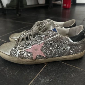 Glittriga sneakers från Golden Goose - Snygga glittriga sneakers från Golden Goose med en rosa stjärna på sidan. Skorna har snörning och en svart häl med märkesnamnet. 