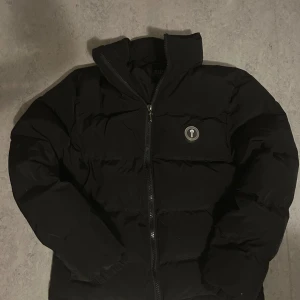 Svart pufferjacka från Trapstar - Snygg svart pufferjacka från Trapstar med dragkedja och logga på bröstet. Jackan har en cool broderad text på ryggen och är perfekt för kyliga dagar. Den har långa ärmar och en hög krage för extra värme.